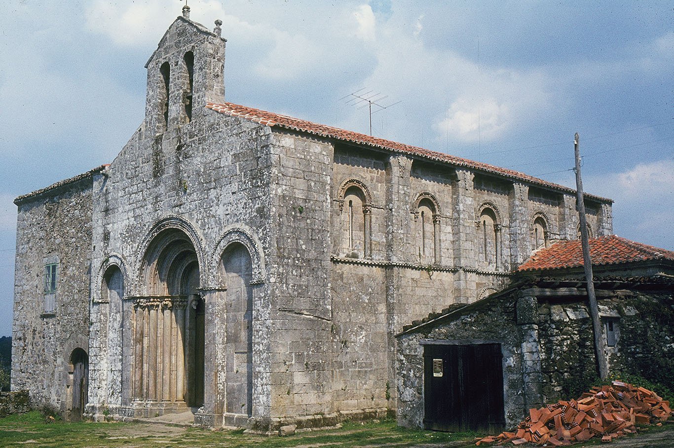 A view of San Paio de Diomondi from the southwest. The deeply recessed portal on the west façade has four orders of moulded arches supported by columns. The plain tympanum rests on corbels. The doorway is flanked by two blind arches. The façade is topped by a simple bellcote with two lights. On the south side, the bays are separated by buttresses. One can see the springing of arches at the top which were never completed. Each bay has a shafted window with a moulded arch. Attached to the left of the church, we can see the rectory. Its rubble masonry contrasts with the fine ashlar of the church. At the right side of the picture, a pile of red roof tiles, linked to repairs on a large shed, adds a dash of color, besides the greenery in front of the church and the blue-grey clouds.
