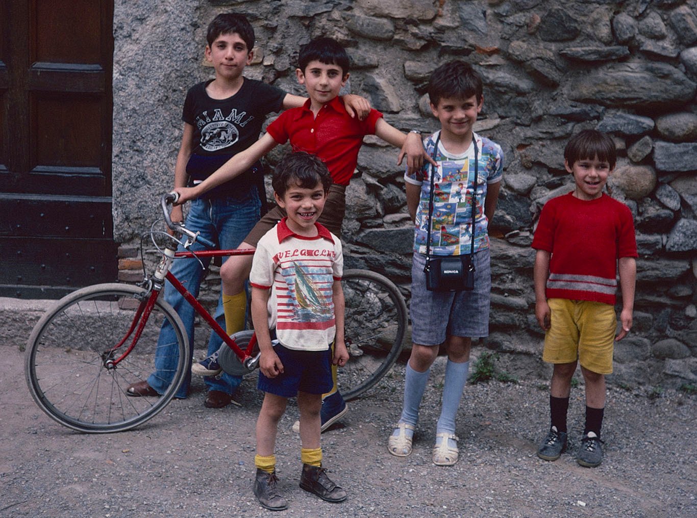 Against a stone wall and doorway, five small boys, one with a bicycle, pose for a picture. They wear colorful tee shirts and, mostly, short pants. One has my Konica camera case around his neck.