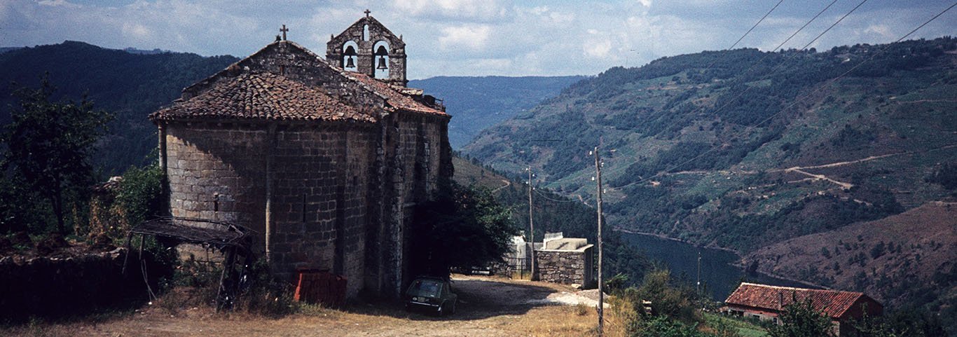 The view shows the semicircular apse of the church, built of well-cut ashlar masonry and covered with a red tile roof. The east wall of the nave is higher than the apse. One can see the bellcote over the west façade with bells in the two openings and a smaller opening in the gable. Beyond the church, the steep, wooded and terraced hills slope down to the Miño river which is visible in the lower right.