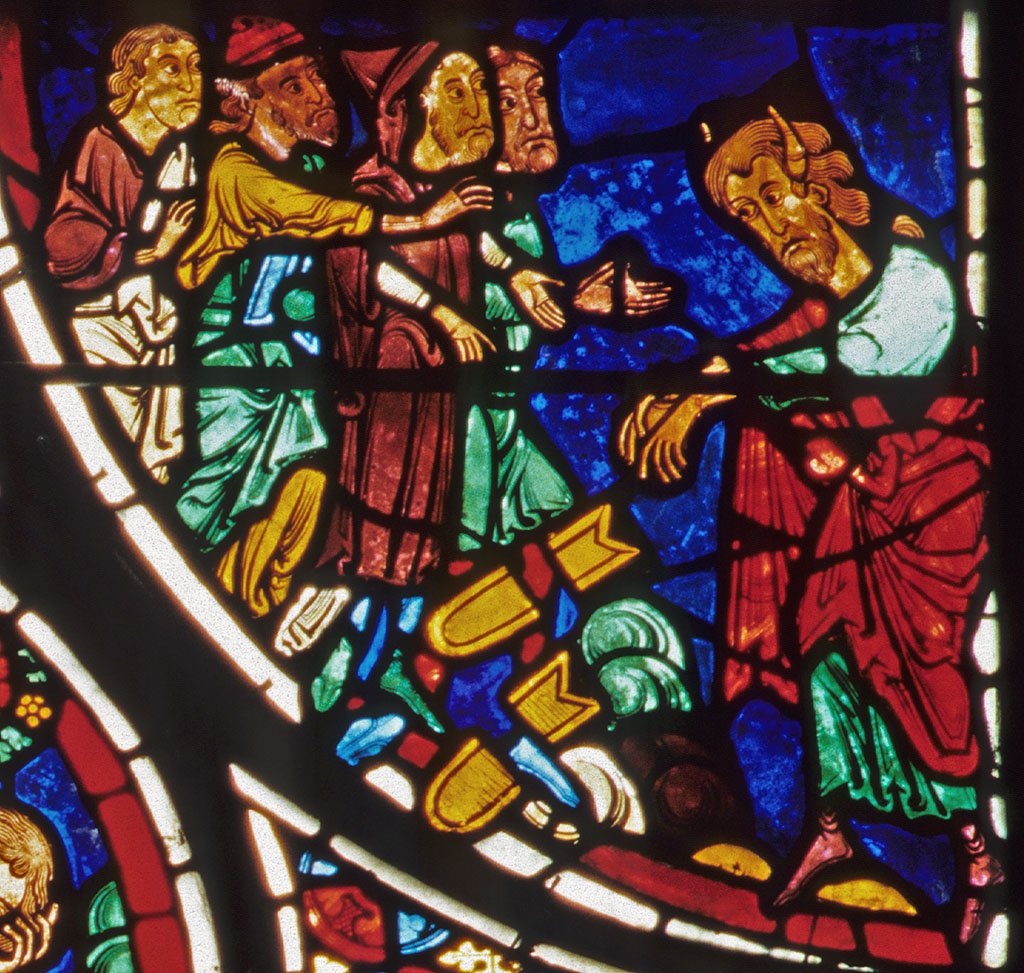 A brightly colored detail of a stained glass window with deep reds and blues, light greens, whites, and golden hues. A horned Moses throws down and shatters the Tablets of the Law in front of four Israelites who gesture at him.