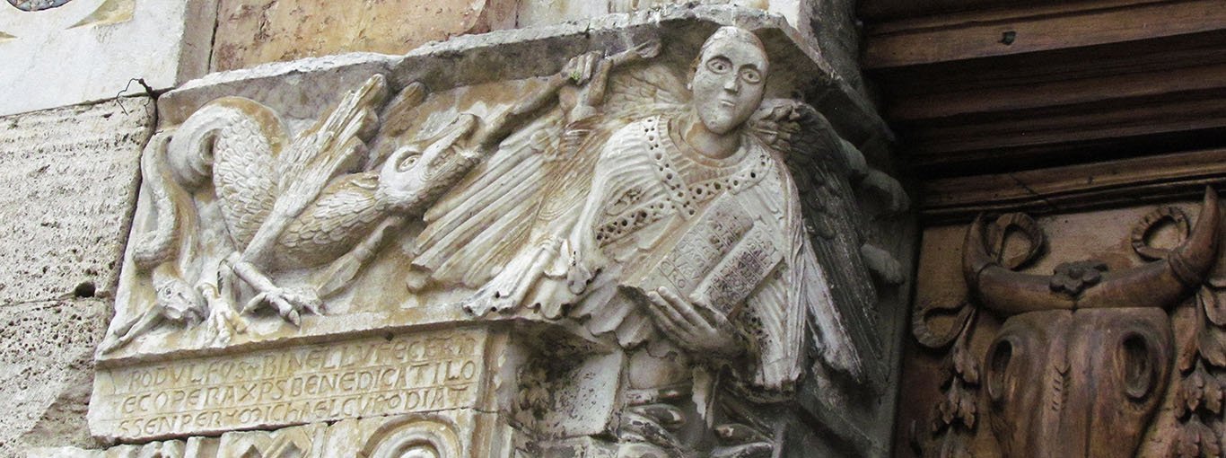 A large rectangular relief at the top of the left door jamb shows the Archangel Michael at the corner, holding an open book with a lengthy inscription. His robes and wings are detailed with fine carving. On the outer face of the piece, we see a winged dragon with talons being speared by St Michael. Below there is a three-line inscription naming the artists with a prayer.
