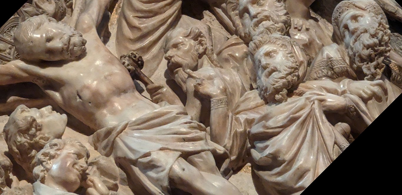 An oblique view of a detail of the Crucifixion scene on the Siena Cathedral pulpit. We see Jesus's head and torso with the deep wound in his side. One man, profiled against the dark shadow of Jesus's body, stares intently at Jesus's slumped face. At the edge of the photo, St. John's head is bent with an expression of grief. To the right of Christ, a balding, bearded man with sharp features and wavy hair over his ears raises a rod with a sponge towards Jesus: his face is almost demonic. To the right, three of the Jewish elders or priests cringe or fall backwards. One points towards Jesus. They are recognizable by their long curly beards and fringed shawls.