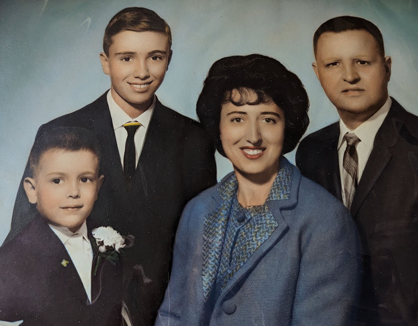 This is a studio photograph against a plain bluish background. My mother and I are in the foreground. My brother and my father are behind us, to each side of my mother. We are formally dressed. My mom has a blue buttoned jacket. My father, my brother, and I have dark suits and ties. I have a white tie and a buttonhole white carnation for my first Holy Communion.
