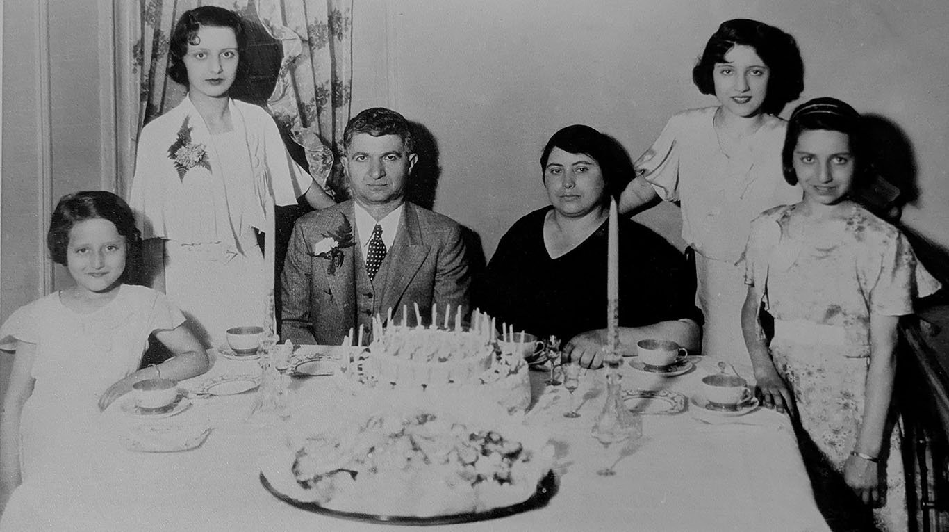 Photograph taken for my grandfather's 50th birthday in 1933. He's wearing a jacket, vest, tie, and a buttonhole flower. Next to him my grandmother is dressed in black. The table has a birthday cake full of candles, a tray of pastries, and a fine set of coffee cups and saucers. Their four daughters (my mother and her three sisters) stand flanking the table.