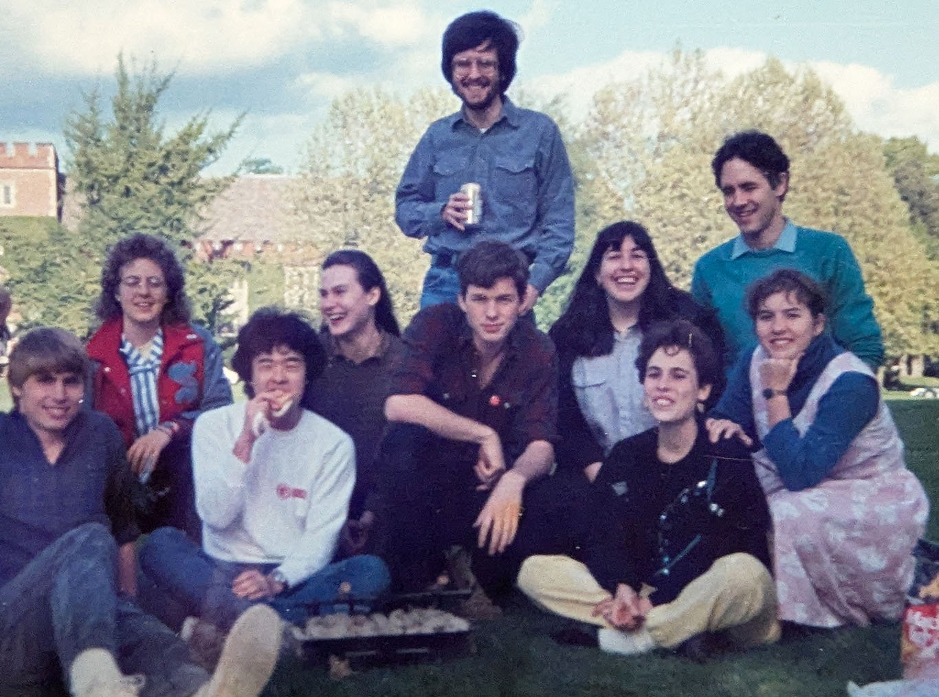 I am standing in a blue shirt and jeans holding a can of soda. In front of me, eight students (three men, five women, and the husband of one student) are sitting or kneeling in two rows on a lawn for the photograph. Behind me, there are trees and the battlemented entrance tower of the Reed College library.