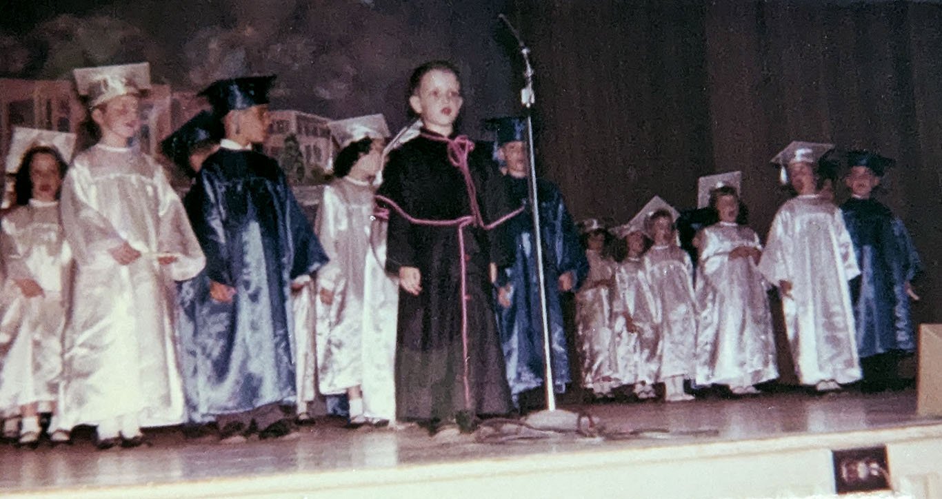 Photograph of my kindergarten graduation ceremony in May 1962. I stand on stage wearing the costume of monsignor in front of a tall microphone. Behind me, the other children stand in rows wearing glossy caps and gowns: the girls are in white, the boys in blue.