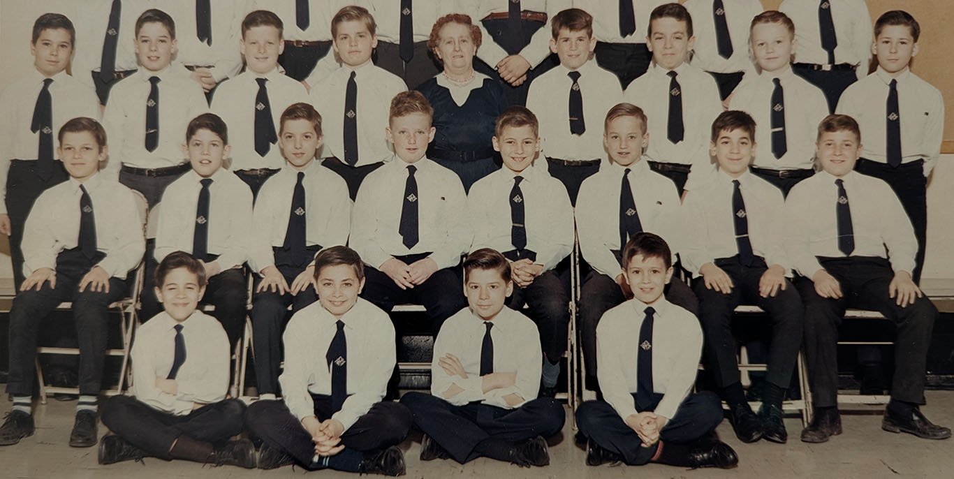 Three rows of boys, four on the floor in the front, eight in a row of chairs behind them, eight standing in the third row and flanking the teacher. All the boys wear white shirts, blue ties with the school emblem and dark trousers.