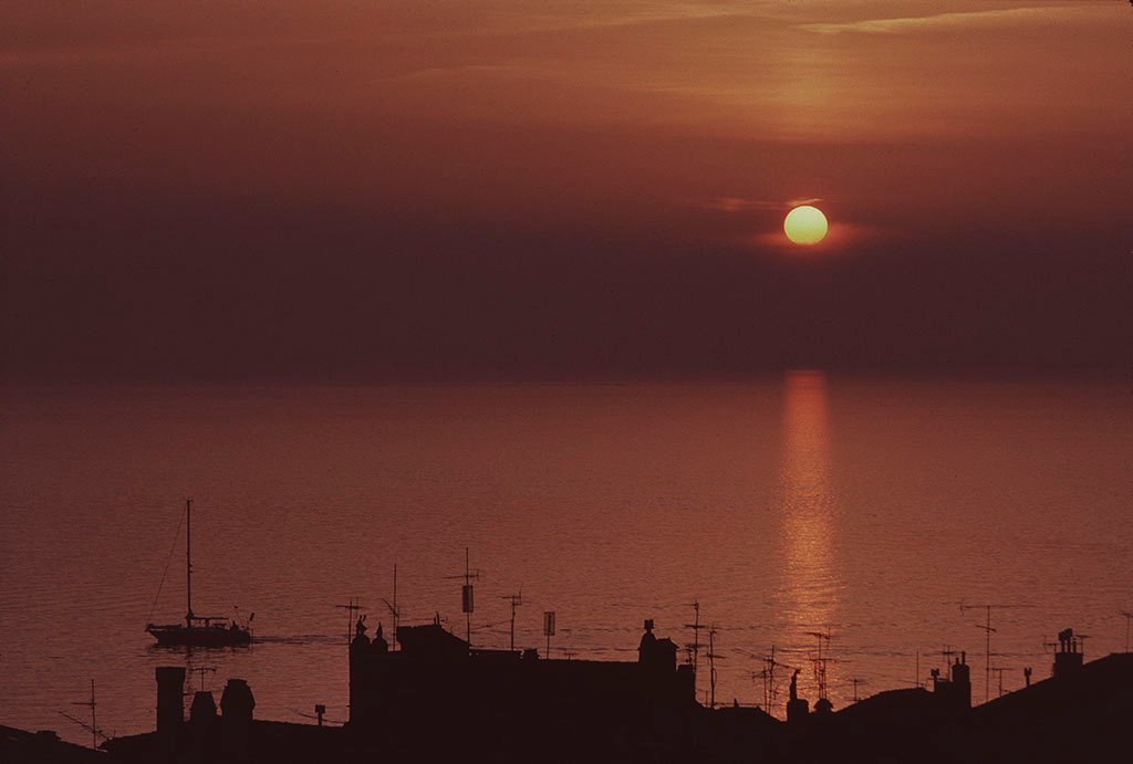 The sun is a golden circle in a deep reddish-orange sky that thickens to a dark reddish grey fog as the sky nears the sea. The water is calm with barely rippling waves. It completely reflects the color of the sky. A long reflected beam of light extends across the surface beneath the setting sun. In the foreground, the black silhouette of roofs, chimneys, and antennas frames the lower part of the picture. Just above, the silhouette of a sailboat, with a tall mast, but no sail, moves towards the left edge.