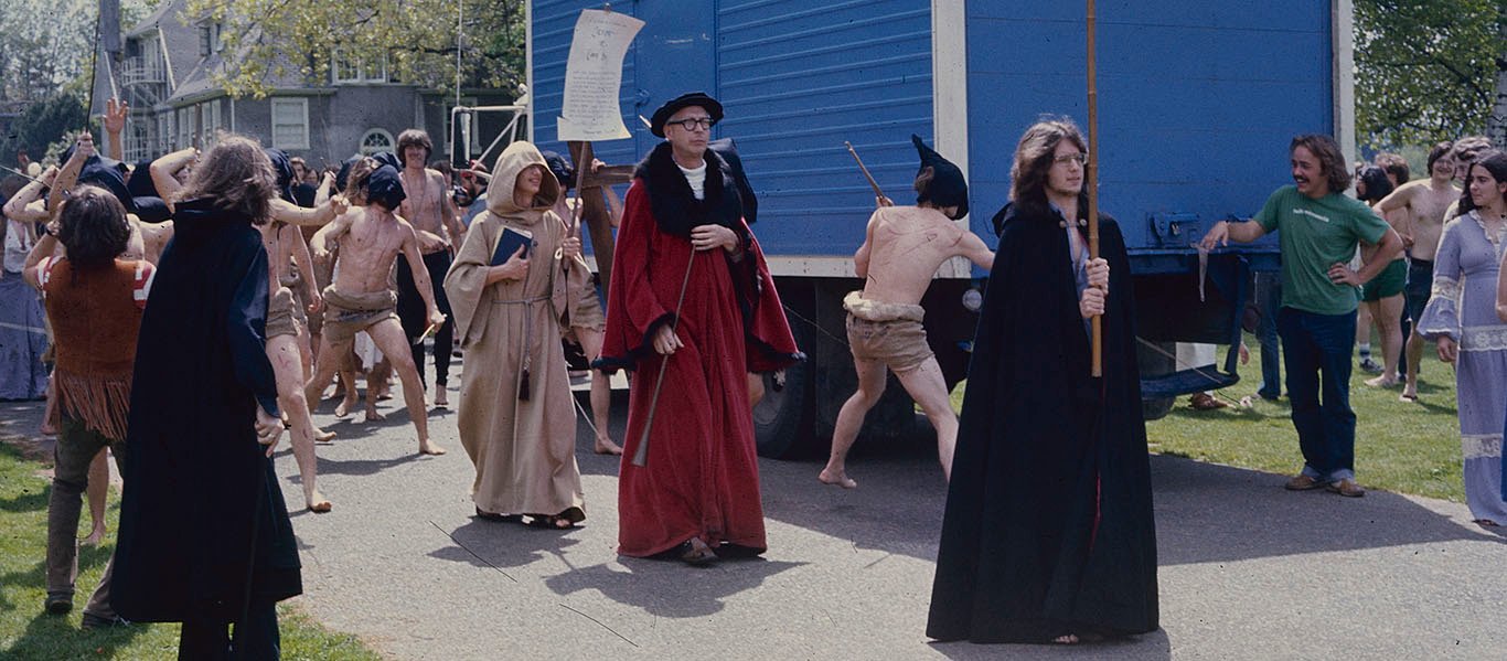 Wearing a black robe and raising a tall processional cross, I am leading a procession of flagellants. Behind me, Professor Hancock walks tall wearing a long red academic robe with a wide black collar and a black hat. Behind him, a student in a hooded burlap robe carries a book and a sign calling for repentance. Further back and around him, all hell breaks loose with male students making demonstrative gestures and whipping themselves with staffs. They are clad only in a burlap cloth wrapped around their waist and have streaks of blood painted on their skin. A large blue truck provides a striking background to highlight the figures and the procession.