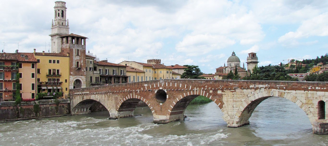 The bridge of brick and white stone spans the swift-flowing Adige river with four wide arches visible in the photograph. The river bank is lined with rows of two, three and four story buildings with yellow and orange pastel colors flanking a towered gate. Beyond the bridge, the domed church and tower of San Giorgio in Braida stand against a blue sky with scattered clouds.